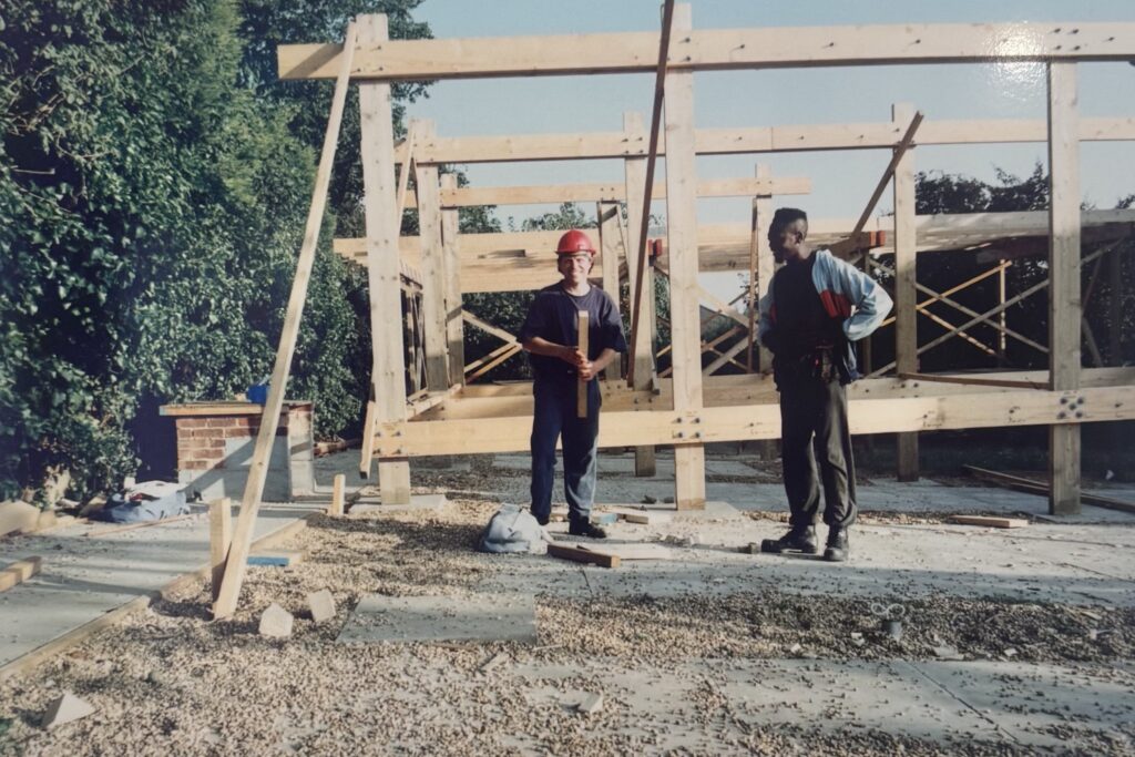 Two men building the frame of a house. Looking to the camera, they are wearing hard hats.